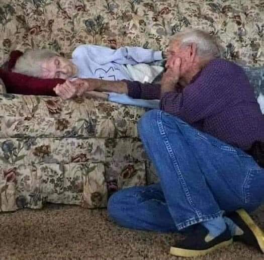 elderly couple holding hands as wife lies on couch